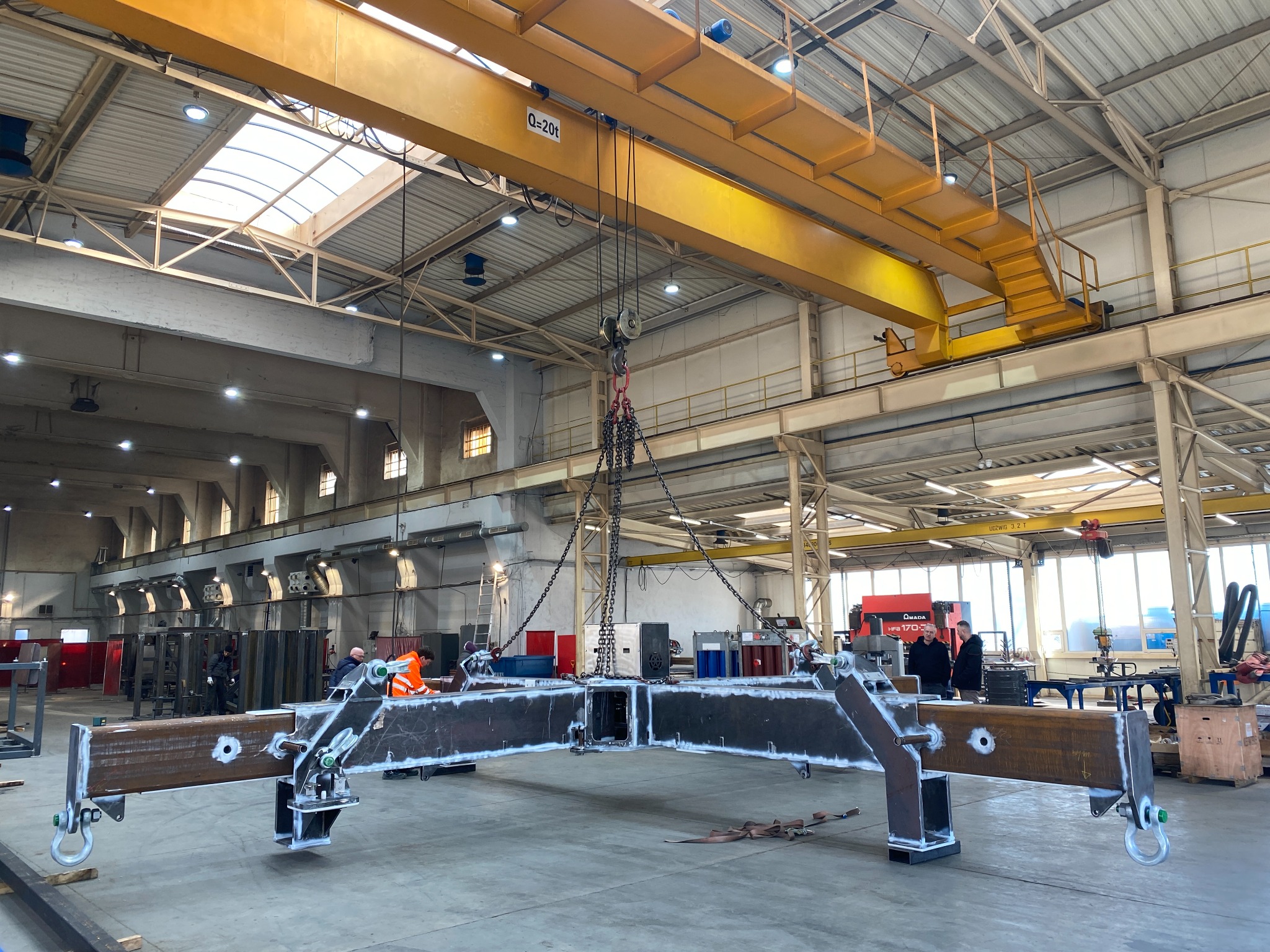 W hali produkcyjnej suwnica podnosi spawany, metalowy element konstrukcyjny w kształcie krzyża. Widoczni pracownicy w odzieży roboczej i spawarka.