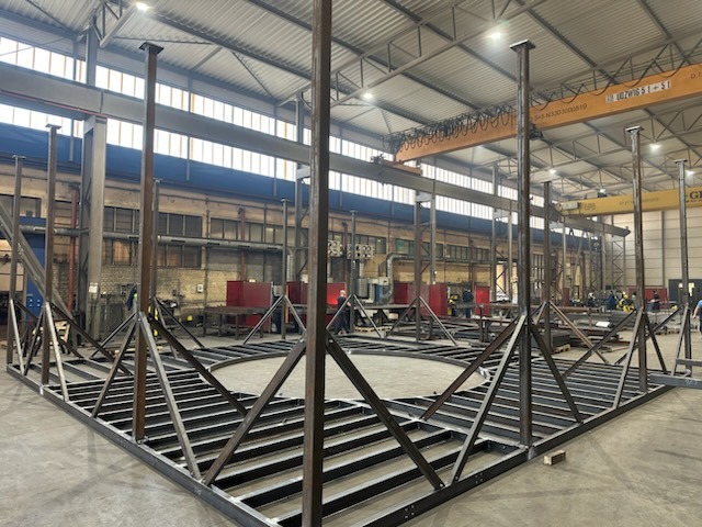 Konstrukcja metalowa z profili w hali produkcyjnej, widoczne spawy, suwnica pod stropem. Elementy rusztowania lub szkieletu budowli w trakcie montażu.