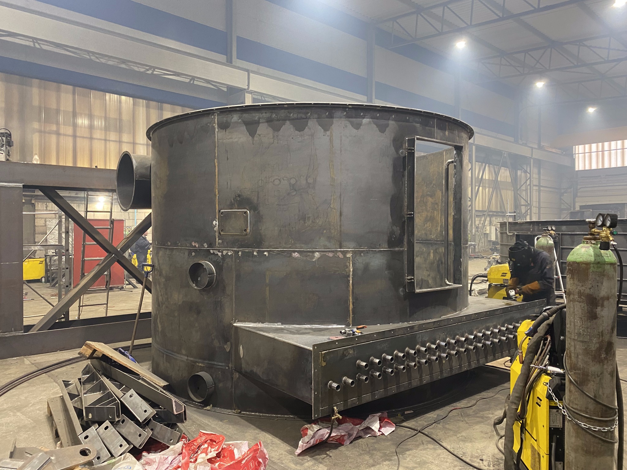 W hali produkcyjnej spawacz łączy elementy dużej, cylindrycznej konstrukcji metalowej z licznymi otworami, używając spawarki. Widoczne butle z gazem i narzędzia.