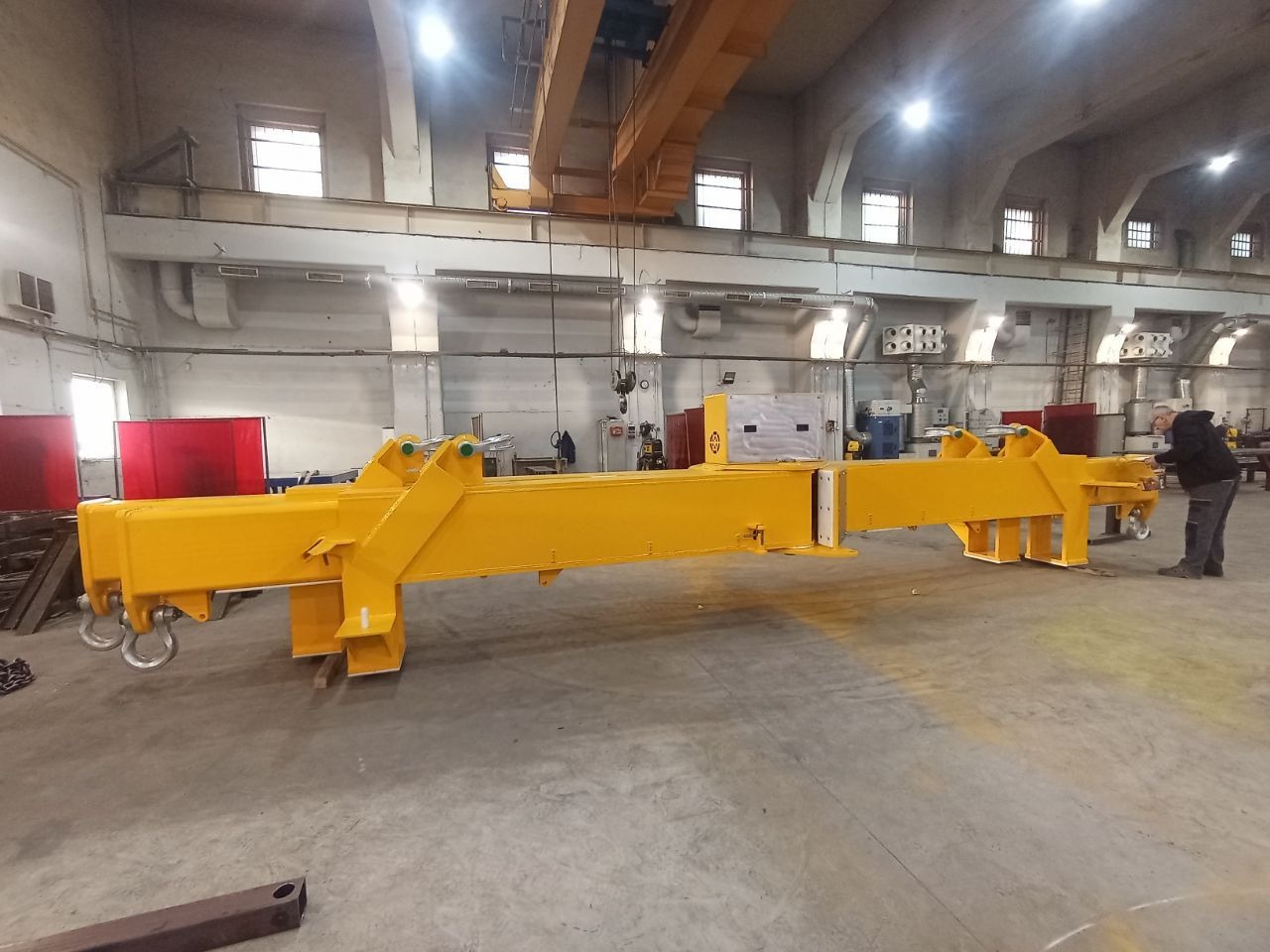 Żółta, pomalowana konstrukcja stalowa w hali produkcyjnej, prawdopodobnie element suwnicy, z widocznymi elementami mocującymi i pracownikiem w tle.