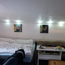 Pokój na poddaszu z ukośnym sufitem, oświetlony szeregiem żarówek, z łóżkiem, stolikiem, kanapą i obrazami na ścianach.