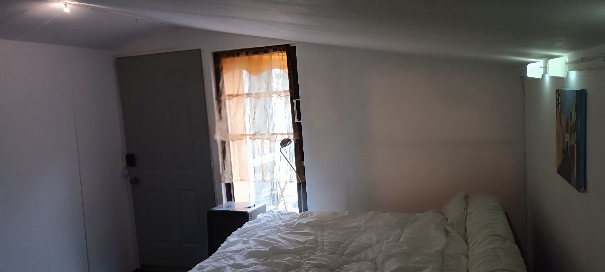 Pokój z łóżkiem przy ścianie, obok okna z beżowymi zasłonami i lampką, widoczny obraz na ścianie i drzwi po lewej stronie