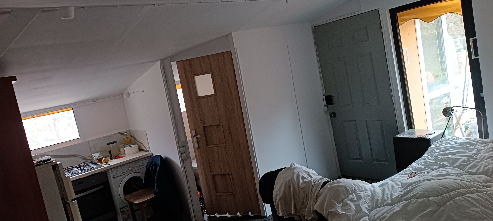 Wnętrze pokoju z aneksem kuchennym, widoczna pralka i kuchenka, drzwi wewnętrzne z drewnopodobną okleiną, oraz drzwi wejściowe z widokiem na zewnątrz.