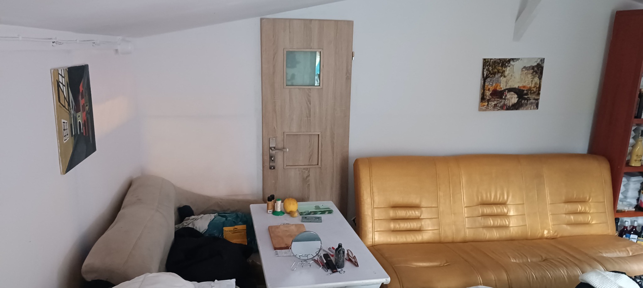 Wnętrze pokoju z beżową sofą, drewnianymi drzwiami z małym okienkiem, stolikiem z lusterkiem i kosmetykami oraz obrazami na ścianach.