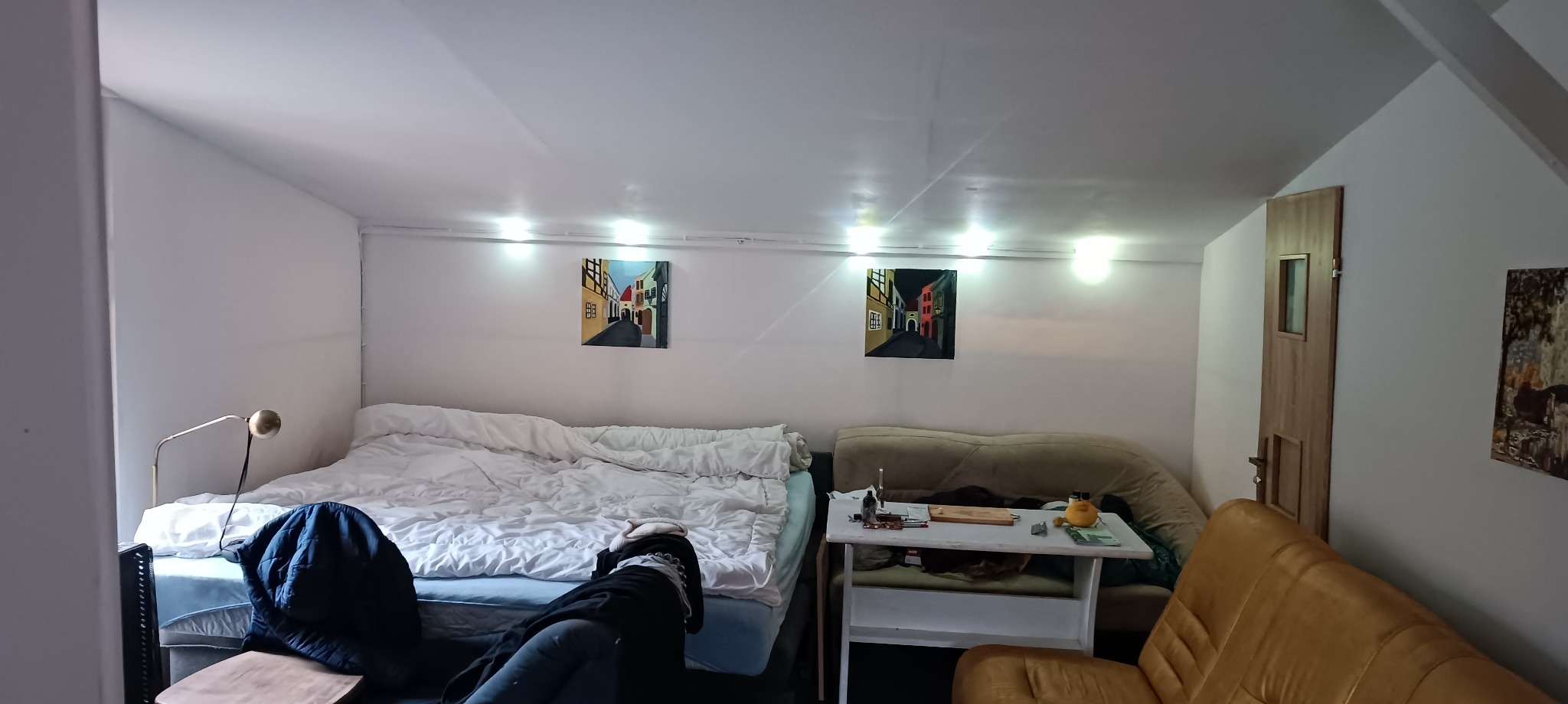 Pokój na poddaszu z ukośnym sufitem, oświetlony szeregiem żarówek, z łóżkiem, stolikiem, kanapą i obrazami na ścianach.