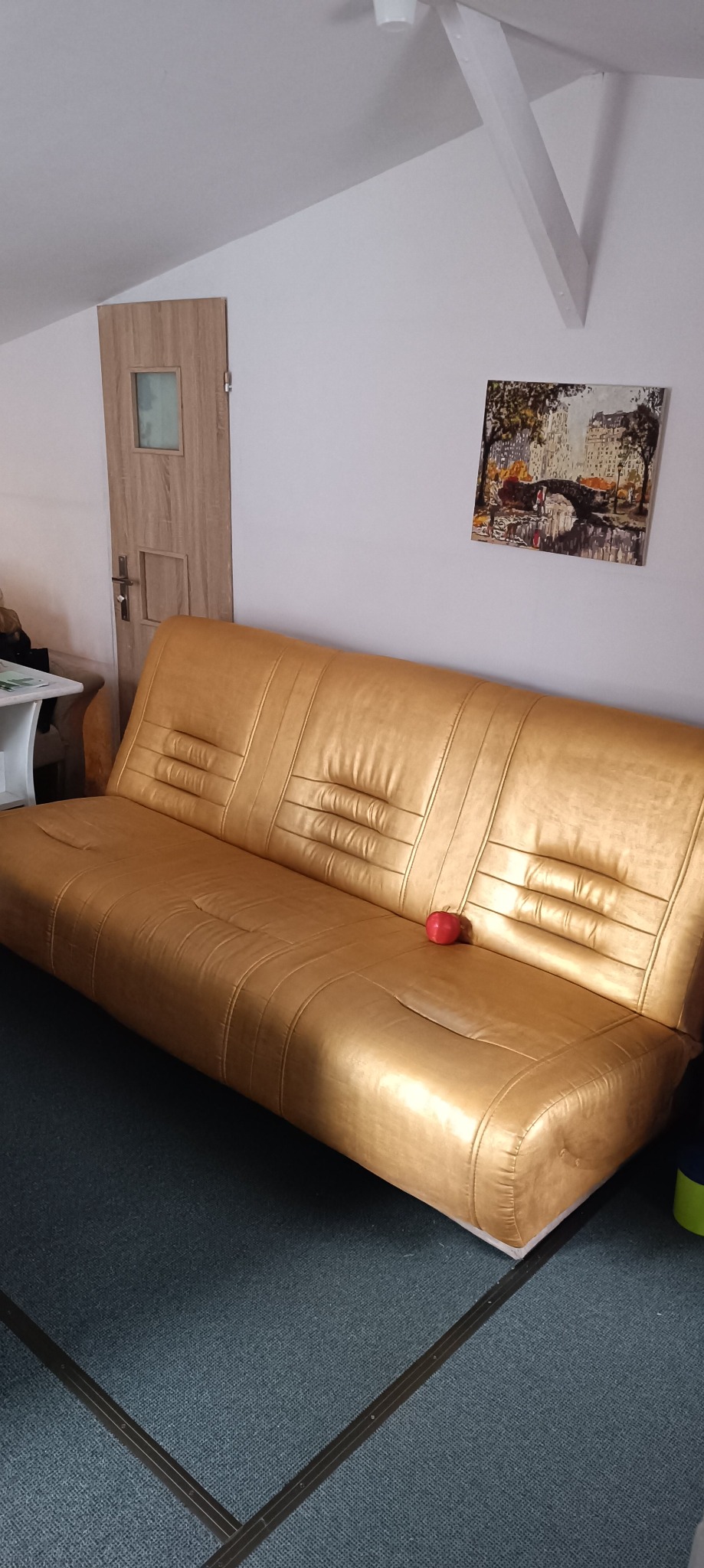 Złota kanapa z czerwoną dekoracyjną jabłkiem na tle ściany z obrazem i drewnianymi drzwiami w pokoju na poddaszu.