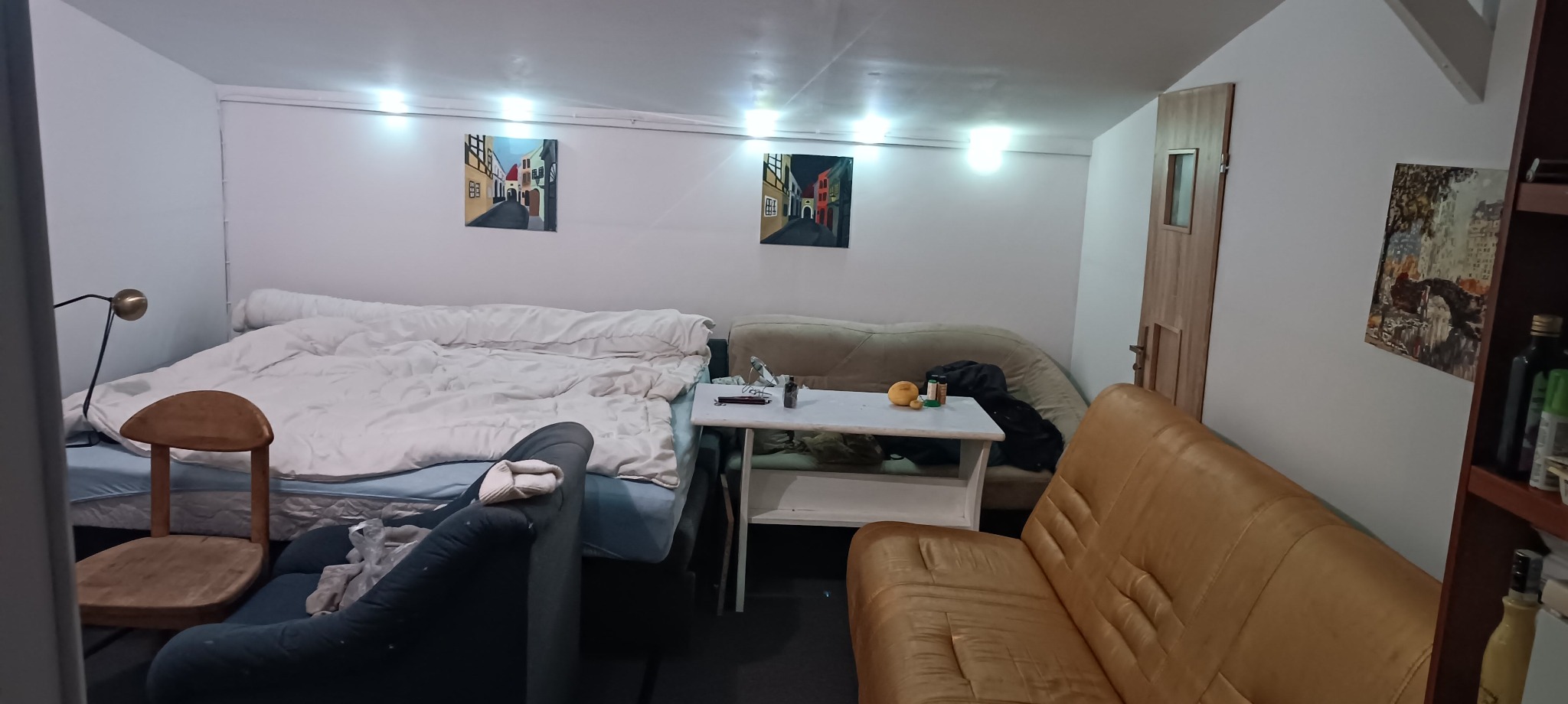 Nieduże pokój na poddaszu ze skośnym sufitem, wyposażony w łóżko, kanapy, stolik i regał, oświetlony listwą LED i lampką biurkową, dekorowany dwoma obrazami z motywem miejskim.