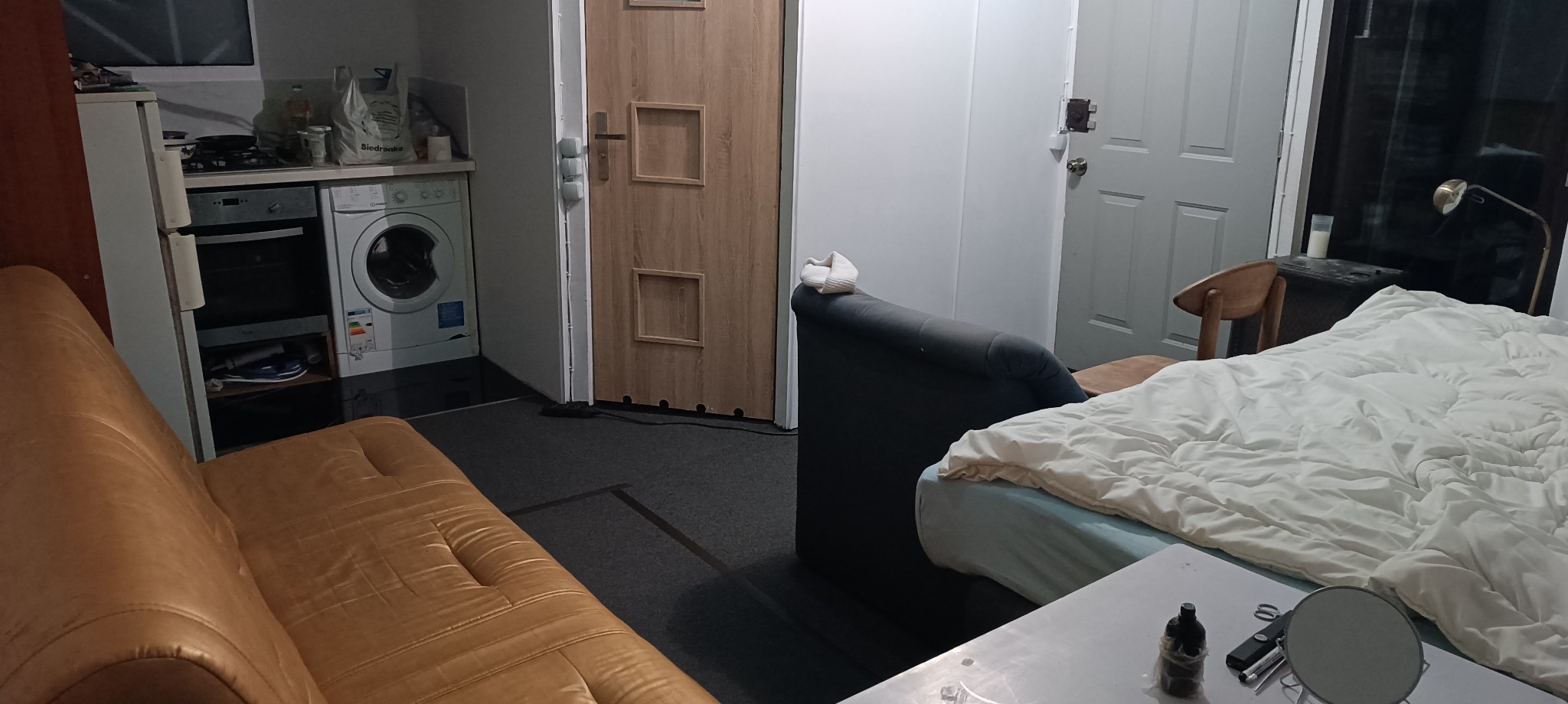 Wnętrze małego mieszkania typu studio z widoczną pralką obok kuchenki, brązową sofą i łóżkiem z pościelą.