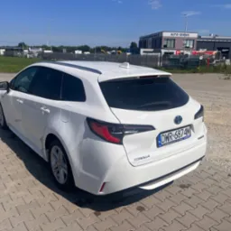 Biała Toyota Corolla kombi, widok z tyłu, zaparkowana na brukowanym placu, z widocznymi kroplami wody na karoserii, tablica rejestracyjna DWR 6874M, w tle budynek auto serwisu.