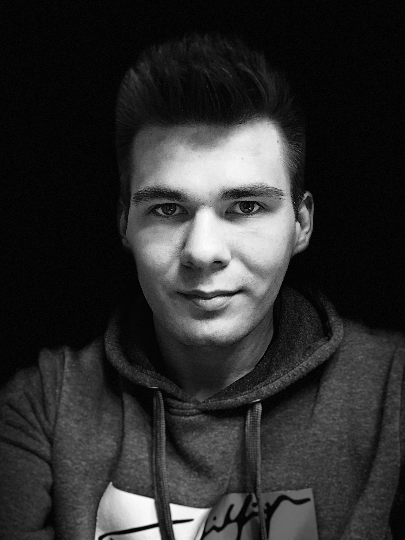 Portret młodego mężczyzny w bluzie z kapturem na czarnym tle, zdjęcie czarno-białe, oświetlenie z przodu.