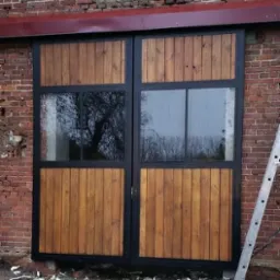 Dwuskrzydłowe drzwi z drewna i szkła w czarnej ramie, zamontowane w murze z czerwonej cegły, drabina oparta o ścianę, widoczne odbicie drzew w szybach.