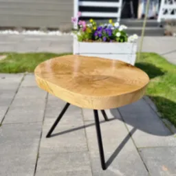 Okrągły, drewniany stolik kawowy z blatem z litego drewna i czarnymi, metalowymi nogami, ustawiony na szarym bruku przed domem z białymi akcentami, w tle donica z kolorowymi kwiatami.