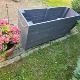 Szara, drewniana skrzynia na rośliny z łańcuchem ozdobnym, stojąca na trawie obok donicy z różowymi hortensjami i kostki brukowej.