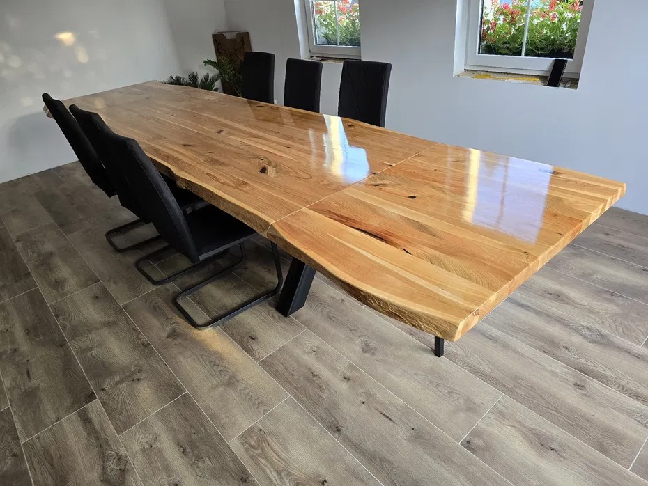 Długi, lakierowany stół z naturalnego drewna o nieregularnych krawędziach, wsparty na metalowych nogach, otoczony czarnymi krzesłami w Gryficach. Widoczne połączenie blatów i refleksy światła...