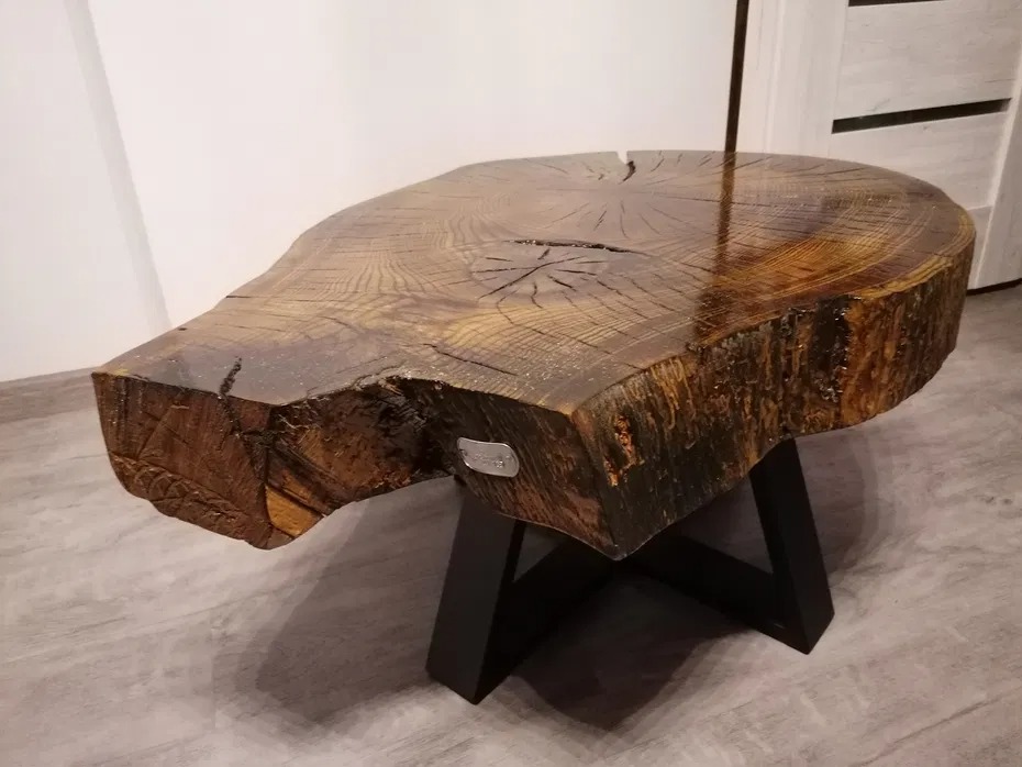 Stolik kawowy z blatem z polerowanego, litego drewna, ukazujący słoje i naturalne pęknięcia, osadzony na czarnej, metalowej podstawie w kształcie litery X.