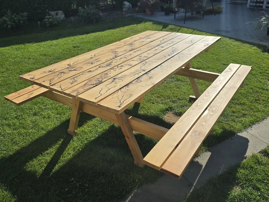 Drewniany stół piknikowy z ławkami, zdobiony wzorem przypominającym rozgałęzienia piorunów, ustawiony na trawniku w ogrodzie.