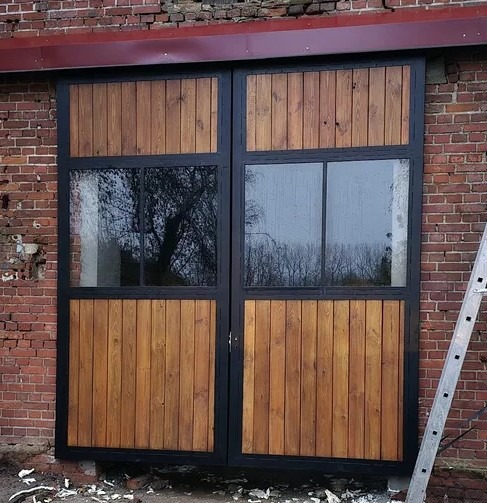 Dwuskrzydłowe drzwi z drewna i szkła w czarnej ramie, zamontowane w murze z czerwonej cegły, drabina oparta o ścianę, widoczne odbicie drzew w szybach.