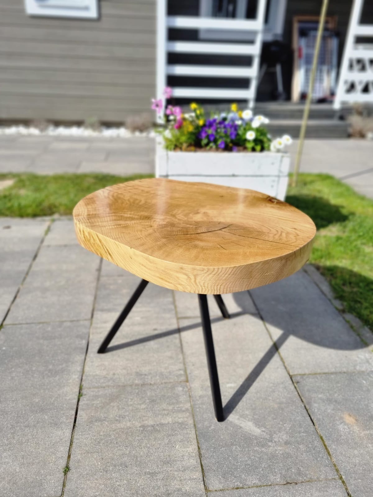 Okrągły, drewniany stolik kawowy z blatem z litego drewna i czarnymi, metalowymi nogami, ustawiony na szarym bruku przed domem z białymi akcentami, w tle donica z kolorowymi kwiatami.