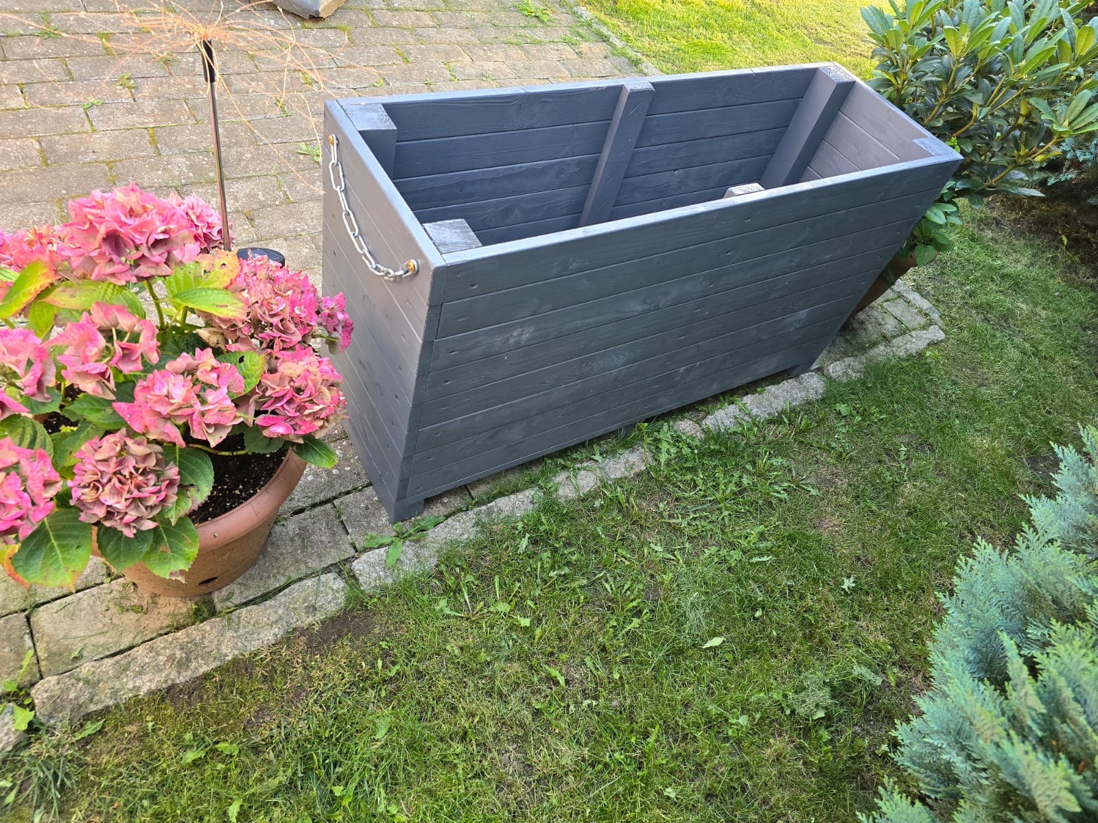 Szara, drewniana skrzynia na rośliny z łańcuchem ozdobnym, stojąca na trawie obok donicy z różowymi hortensjami i kostki brukowej.