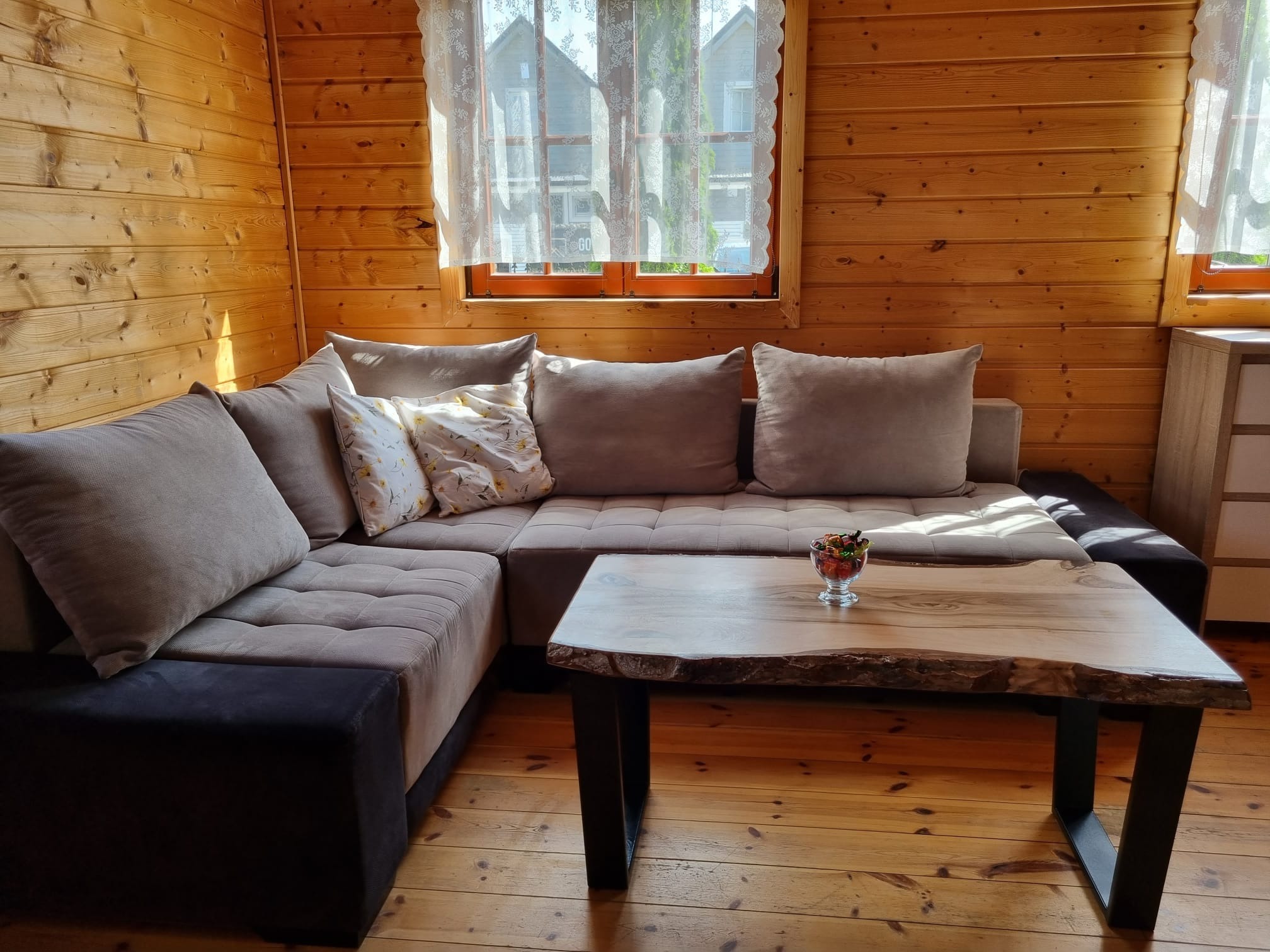 Wnętrze drewnianego domu z narożną sofą w odcieniach beżu, stolikiem kawowym z blatem z surowego drewna i czarnymi nogami, oraz drewnianą podłogą. Na stoliku stoi szklana miseczka z kolorowymi...