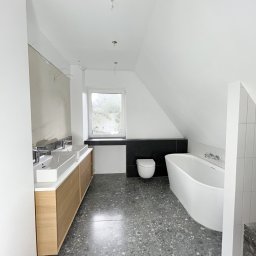 P&O remonty i wykończenia ERYK OSTROWSKI - Łazienka na poddaszu w trakcie wykańczania: dwie umywalki z drewnianą szafką, wolnostojąca wanna, toaleta pod oknem, szara podłoga lastryko, ściany częściowo przygotowane pod malowanie.