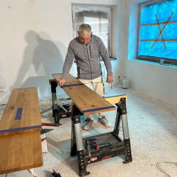 P&O remonty i wykończenia ERYK OSTROWSKI - Stolarz mierzy drewniany blat na kozłach montażowych w pomieszczeniu w trakcie prac wykończeniowych, okna zabezpieczone folią malarską.