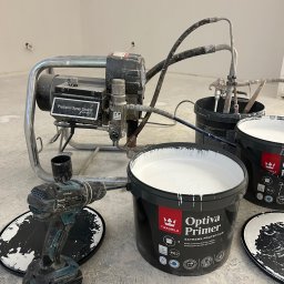 P&O remonty i wykończenia ERYK OSTROWSKI - Sprzęt do natryskowego malowania ścian Tikkurila Optiva Primer, pistolet natryskowy, wiadro z farbą, wiertarko-wkrętarka Makita i pokrywki od wiader na betonowej posadzce w trakcie prac malarskich.