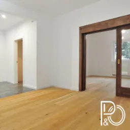 Jasna drewniana podłoga ułożona w nowym mieszkaniu, widoczne białe ściany, przesuwne drzwi z ciemnego drewna oraz logo firmy w rogu zdjęcia.