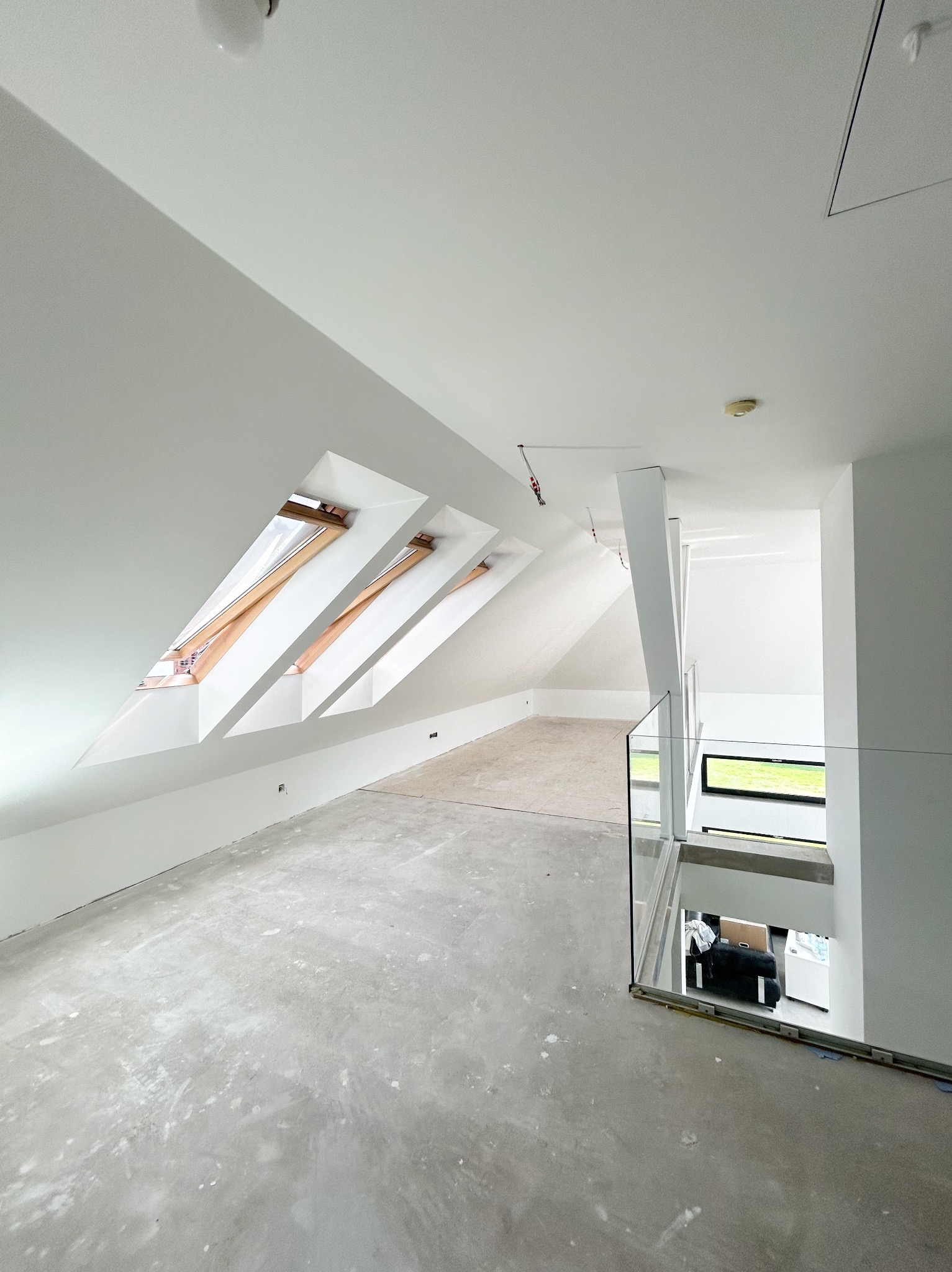 Poddasze w stanie deweloperskim z trzema oknami dachowymi, surową wylewką i szklaną balustradą schodów.
