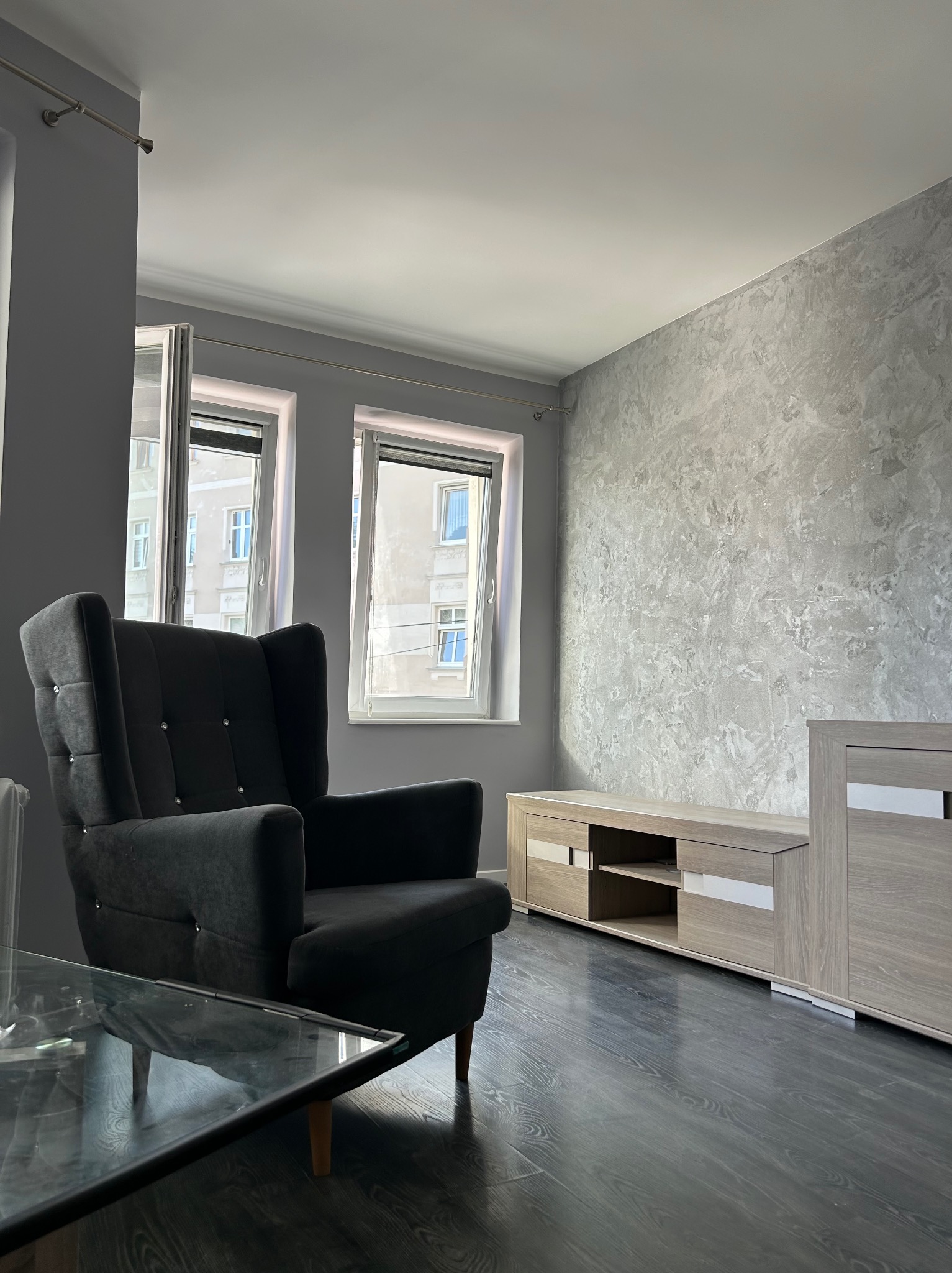 Pokój dzienny w szarościach: dekoracyjna ściana, ciemna podłoga, meble w odcieniach drewna i czarny fotel z kryształkami.