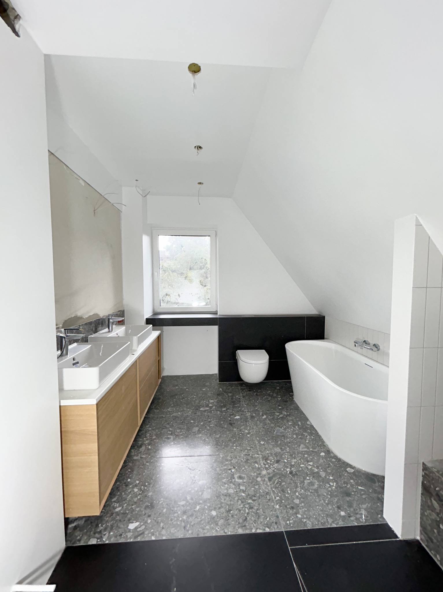 Łazienka na poddaszu w trakcie wykańczania: dwie umywalki z drewnianą szafką, wolnostojąca wanna, toaleta pod oknem, szara podłoga lastryko, ściany częściowo przygotowane pod malowanie.