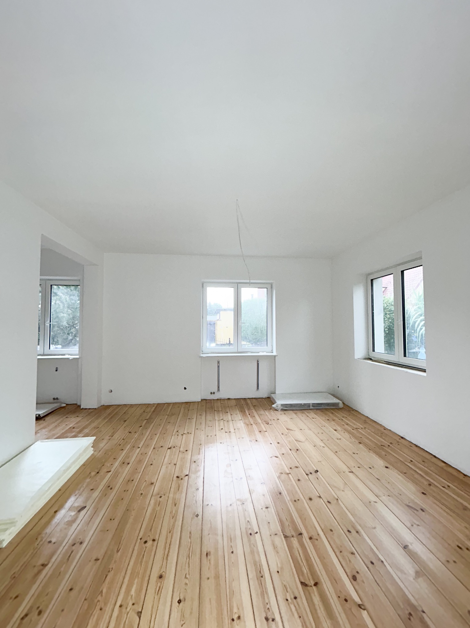 Jasne, świeżo wykończone wnętrze z drewnianą podłogą, białymi ścianami i dwoma oknami; widoczne grzejniki, przewód zwisający z sufitu i płyty styropianu w rogu.