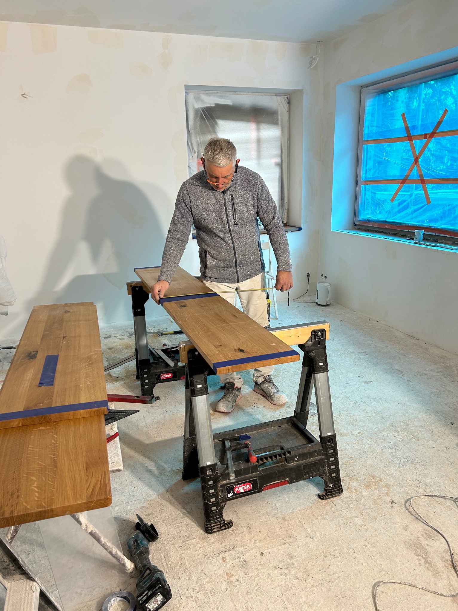 Stolarz mierzy drewniany blat na kozłach montażowych w pomieszczeniu w trakcie prac wykończeniowych, okna zabezpieczone folią malarską.