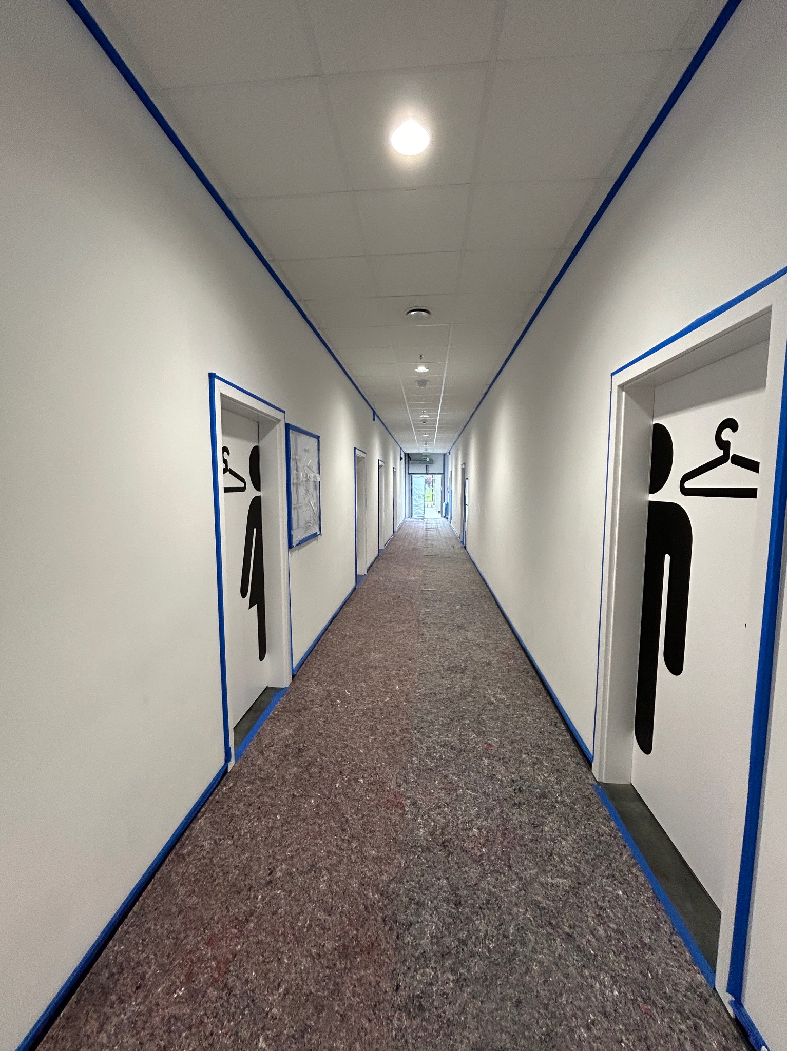 Korytarz w Przecławiu zabezpieczony folią malarską i taśmą ochronną przed malowaniem ścian i ościeżnic, widoczne oznaczenia damskiej i męskiej szatni.