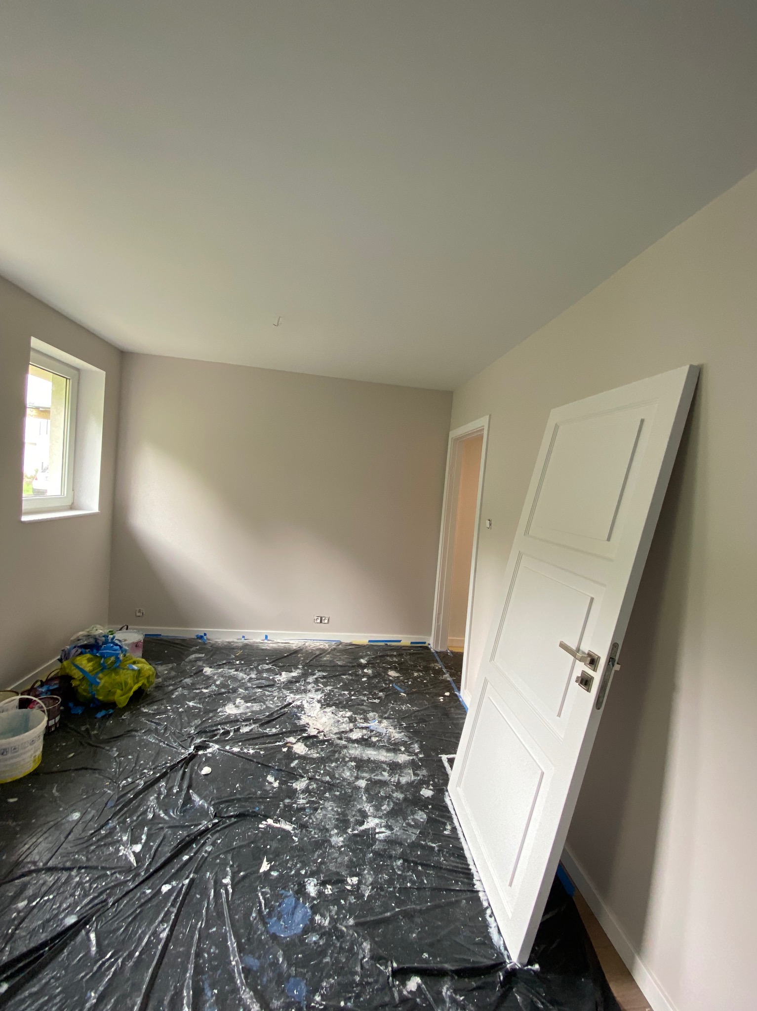 Pokój w trakcie malowania na szaro, podłoga zabezpieczona czarną folią malarską z plamami farby, białe drzwi opierają się o ścianę, widoczne wiadro z farbą i okno.