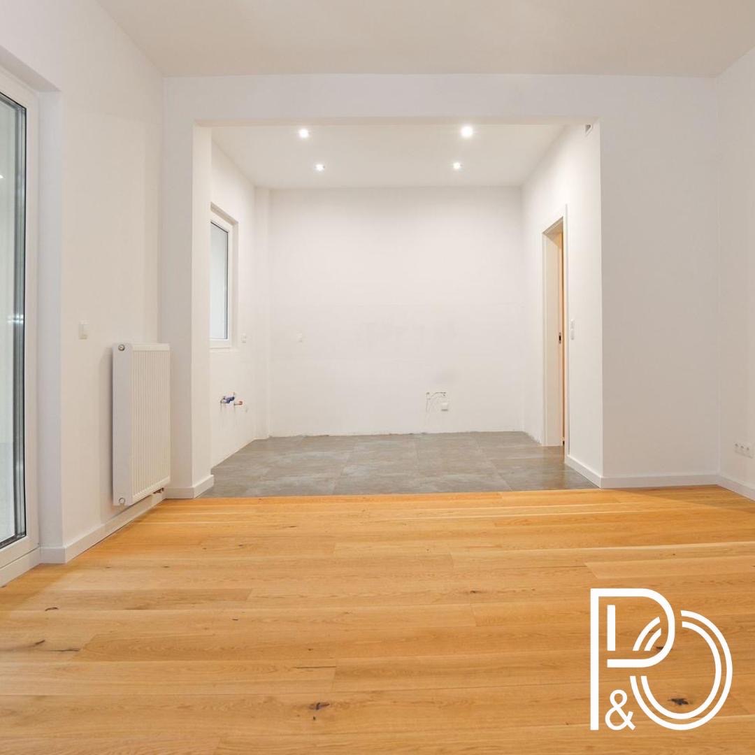 Wnętrze mieszkania w trakcie prac: położona drewniana podłoga, surowa ściana w głębi, widoczny grzejnik i okno, logo firmy w prawym dolnym rogu.