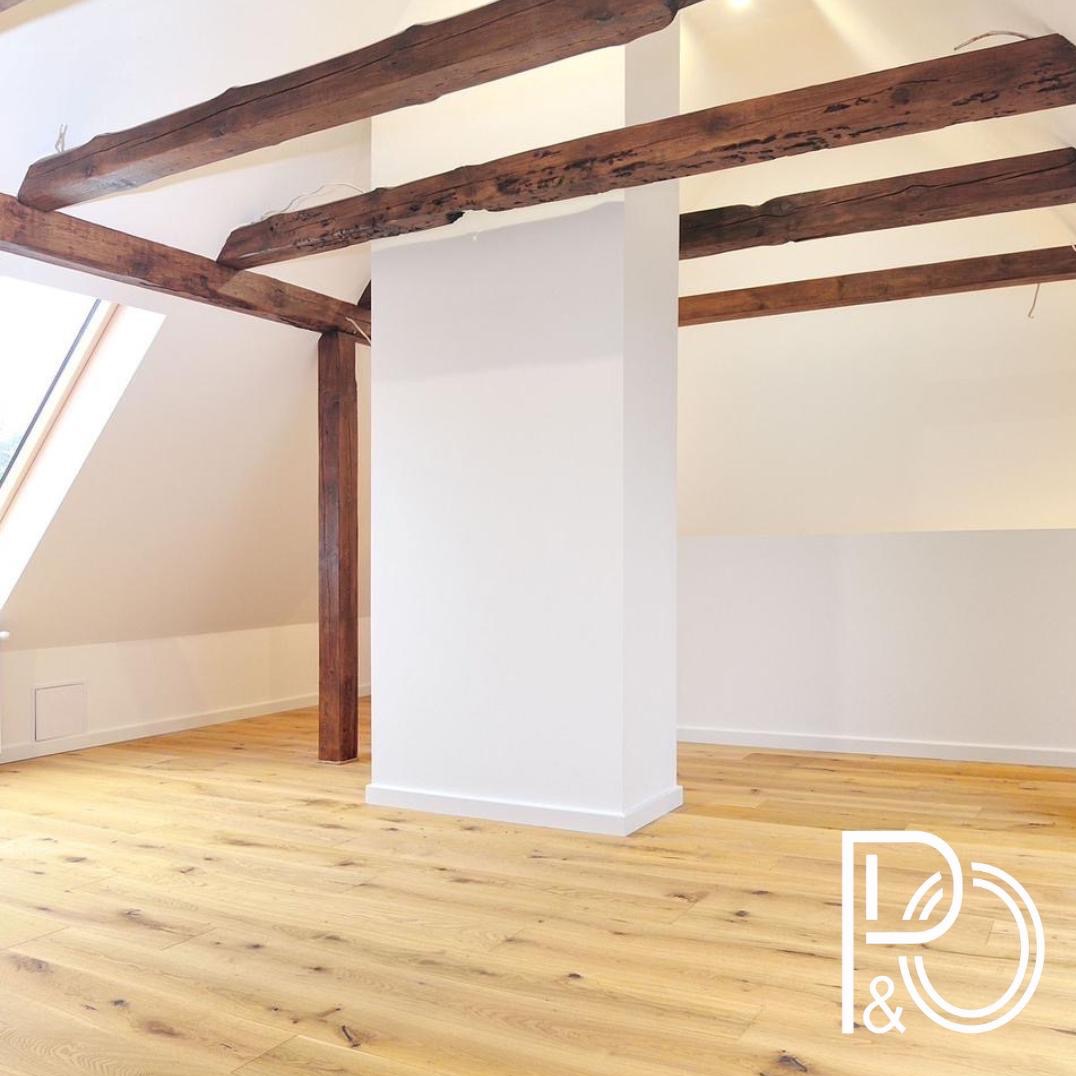 Poddasze po remoncie z drewnianymi belkami stropowymi, jasną podłogą panelową i białymi ścianami, widoczne okno dachowe.