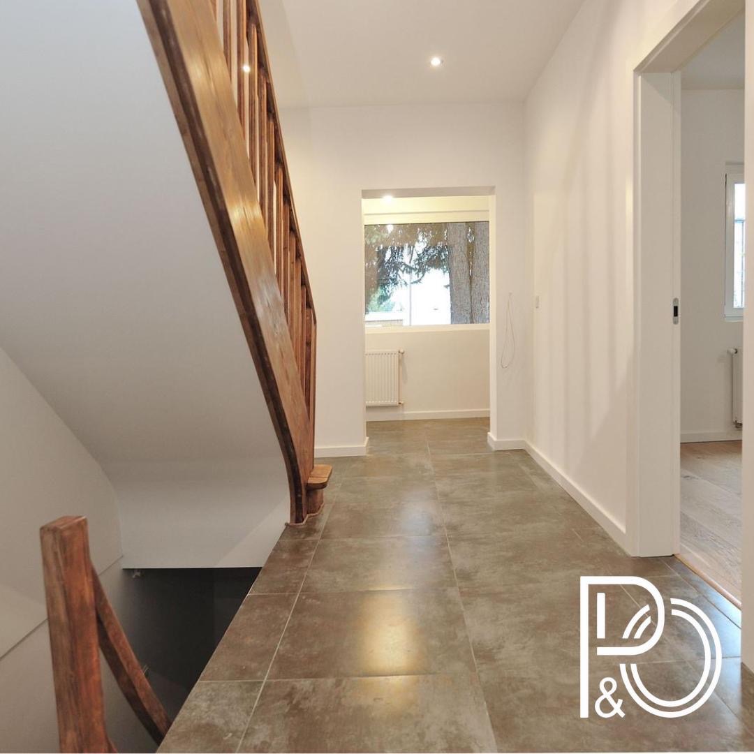 Wnętrze korytarza z brązowymi płytkami podłogowymi, drewnianymi schodami z balustradą i białymi ścianami; widok na okno i drzwi do innego pomieszczenia.