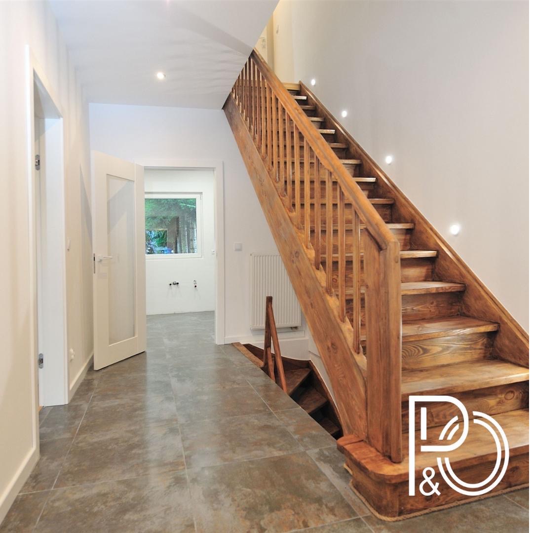 Jasne wnętrze z drewnianymi schodami z balustradą, szarymi płytkami podłogowymi i otwartymi drzwiami do kolejnego pomieszczenia z oknem. Logo firmy w prawym dolnym rogu.