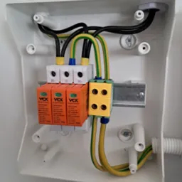 Rozdzielnica elektryczna w białej obudowie z trzema ogranicznikami przepięć VCX T1T2-DC, żółto-zielonymi przewodami uziemiającymi i czarnymi przewodami, zamontowana na szynie DIN.