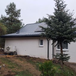 FIRMA USŁUGOWO BUDOWLANA BUDMAR MM SPÓŁKA Z OGRANICZONĄ ODPOWIEDZIALNOŚCIĄ - Dom jednorodzinny z szarym dachem, panelami słonecznymi i zamontowaną pompą ciepła na zewnątrz, otoczony zielenią i drzewami iglastymi w pochmurny dzień.