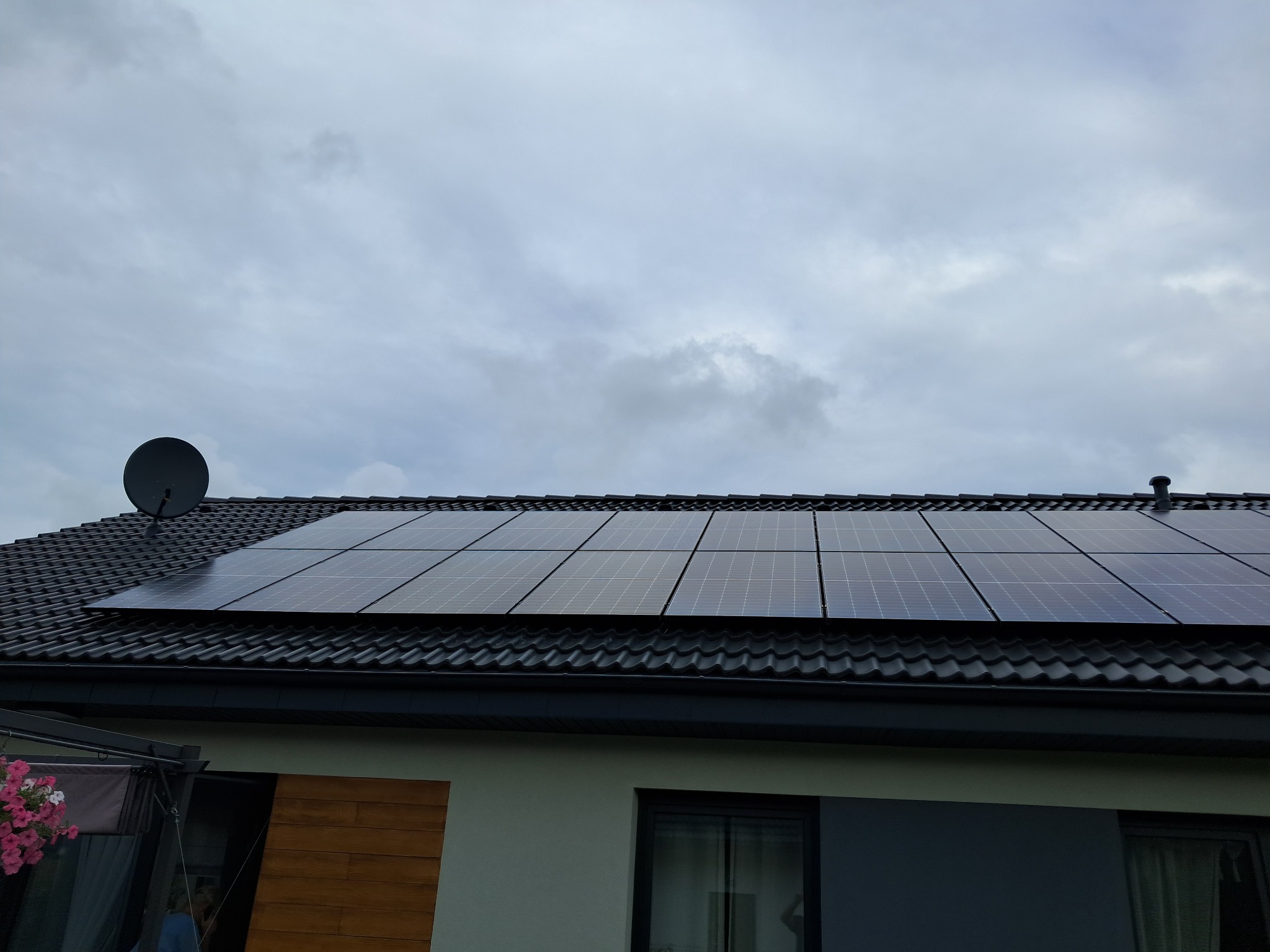 Panele fotowoltaiczne na ciemnym dachu domu jednorodzinnego w Bydgoszczy, widok z dołu na tle pochmurnego nieba. Na dachu widoczna antena satelitarna. Część elewacji domu widoczna w dolnej części...