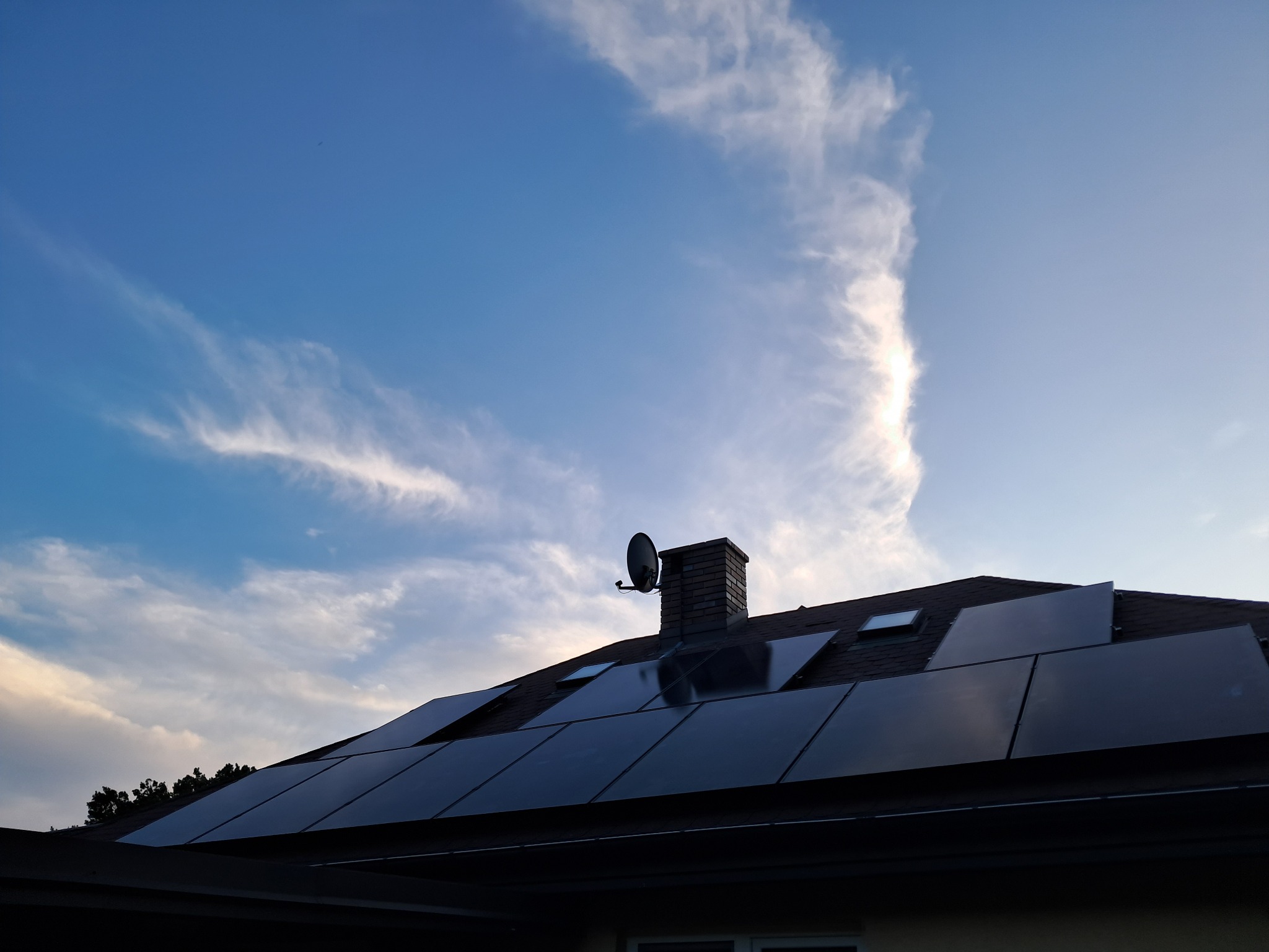Panele słoneczne na dachu domu jednorodzinnego, widok z dołu na tle błękitnego nieba z chmurami, komin z anteną satelitarną.