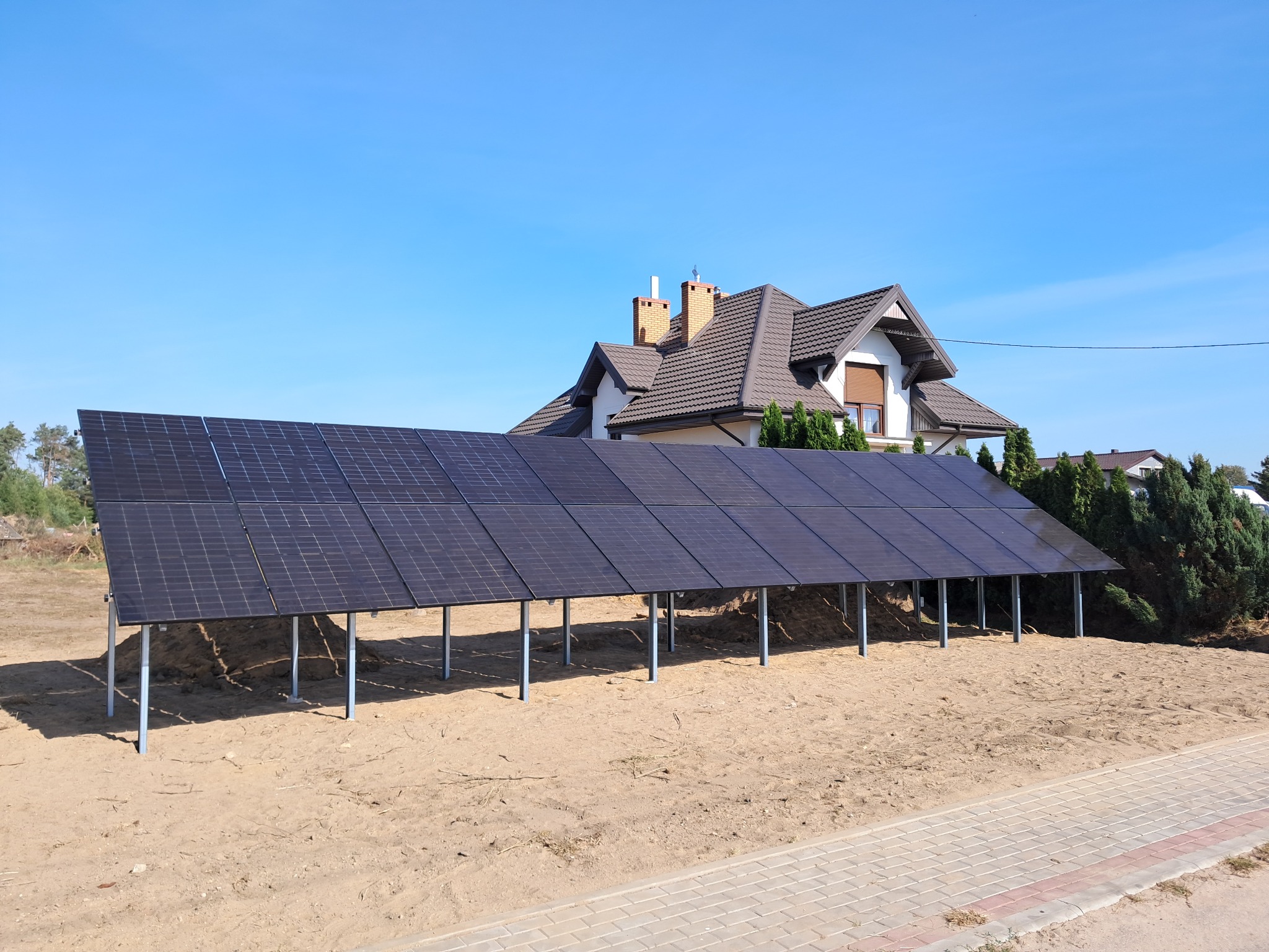 Rząd paneli fotowoltaicznych na metalowych podporach, na piaszczystym podłożu, z widokiem na dom z brązowym dachem i ceglanymi kominami, w słoneczny dzień.