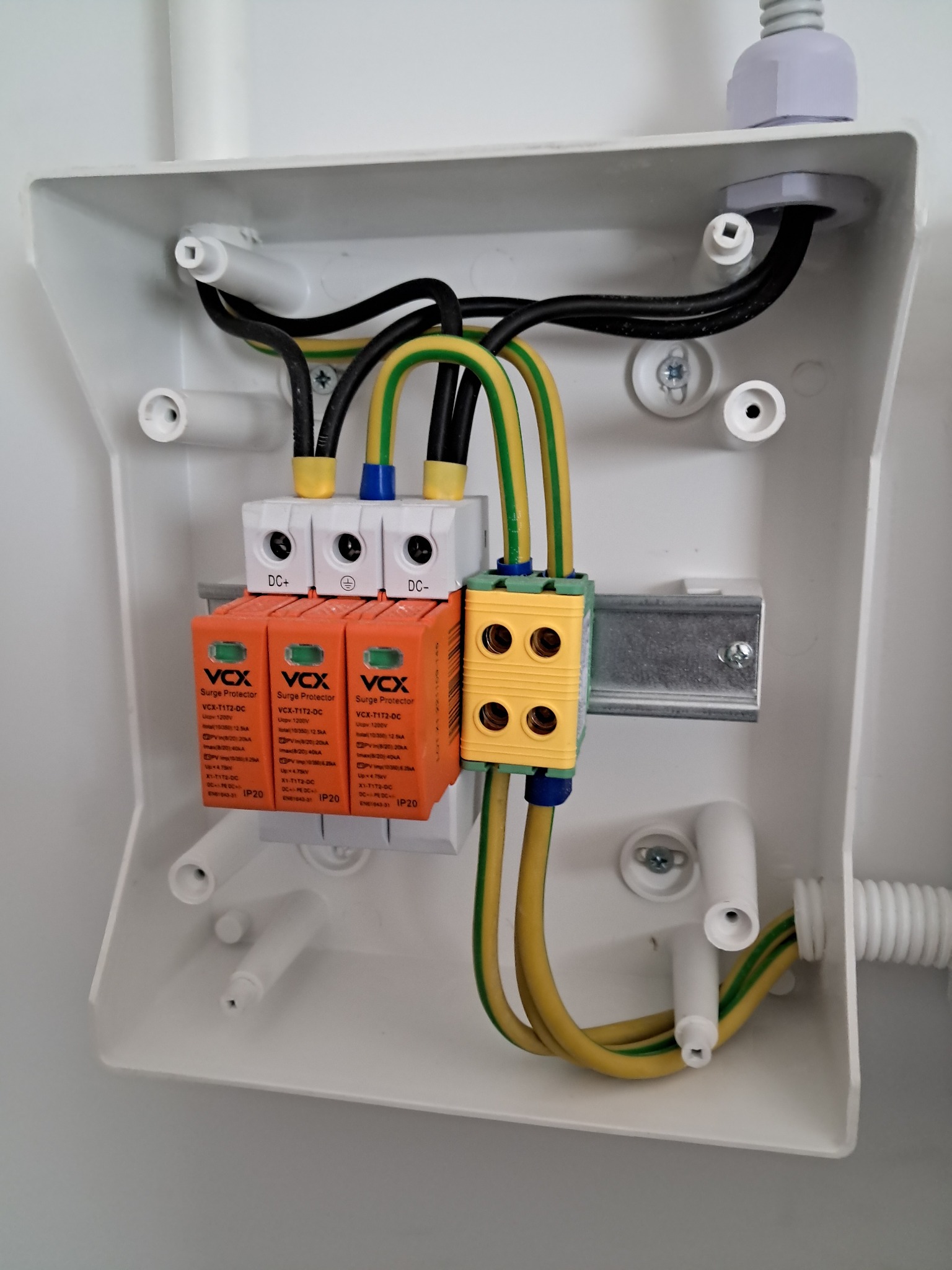 Rozdzielnica elektryczna w białej obudowie z trzema ogranicznikami przepięć VCX T1T2-DC, żółto-zielonymi przewodami uziemiającymi i czarnymi przewodami, zamontowana na szynie DIN.