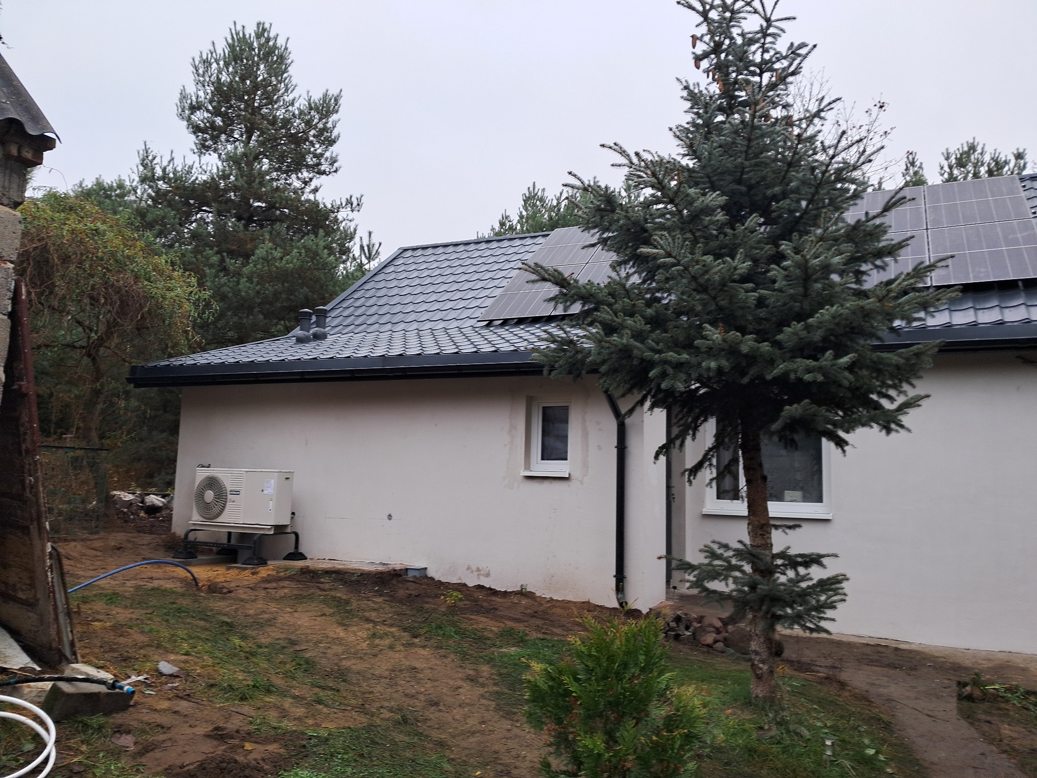 Dom jednorodzinny z szarym dachem, panelami słonecznymi i zamontowaną pompą ciepła na zewnątrz, otoczony zielenią i drzewami iglastymi w pochmurny dzień.