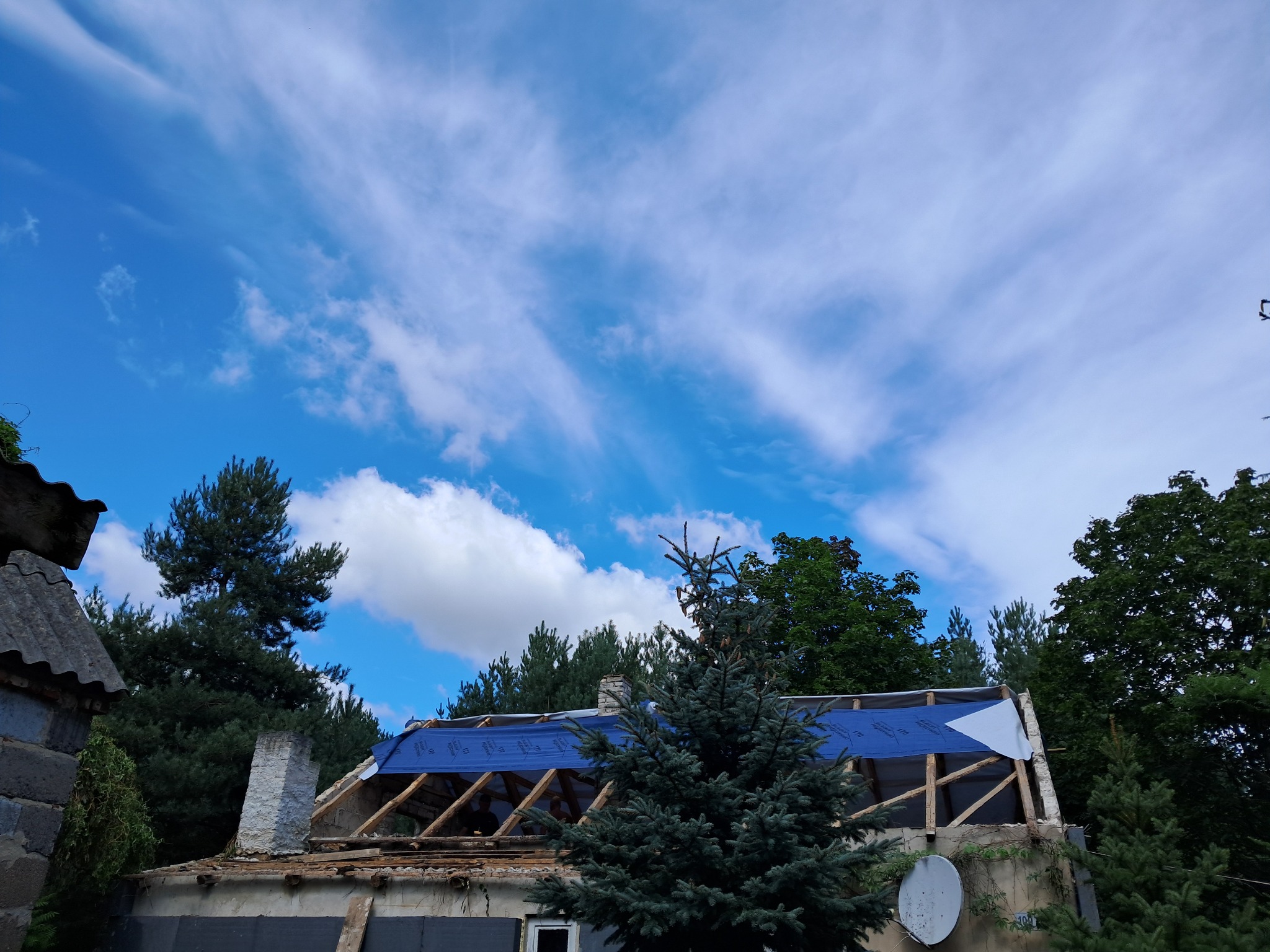 Demontaż dachu z widoczną konstrukcją drewnianą, zabezpieczoną niebieską folią, na tle błękitnego nieba z chmurami i zieleni drzew.