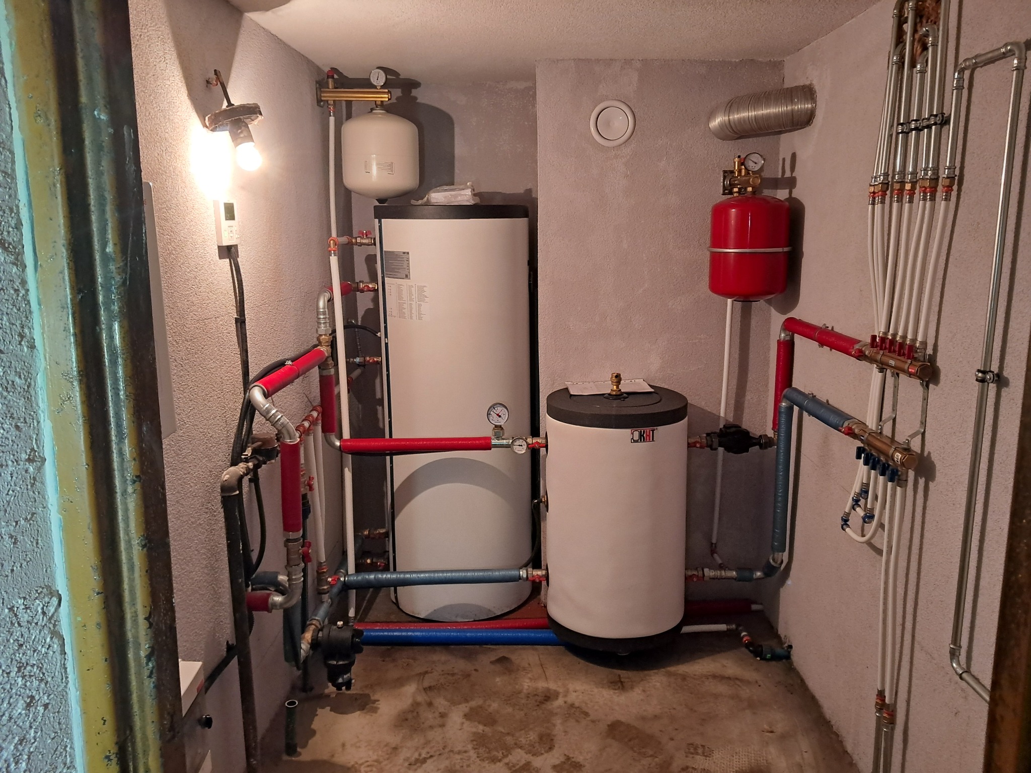 Kotłownia z dwoma zbiornikami, czerwonym naczyniem wzbiorczym, rurami w kolorach czerwonym i niebieskim oraz oświetleniem żarówką na ścianie.
