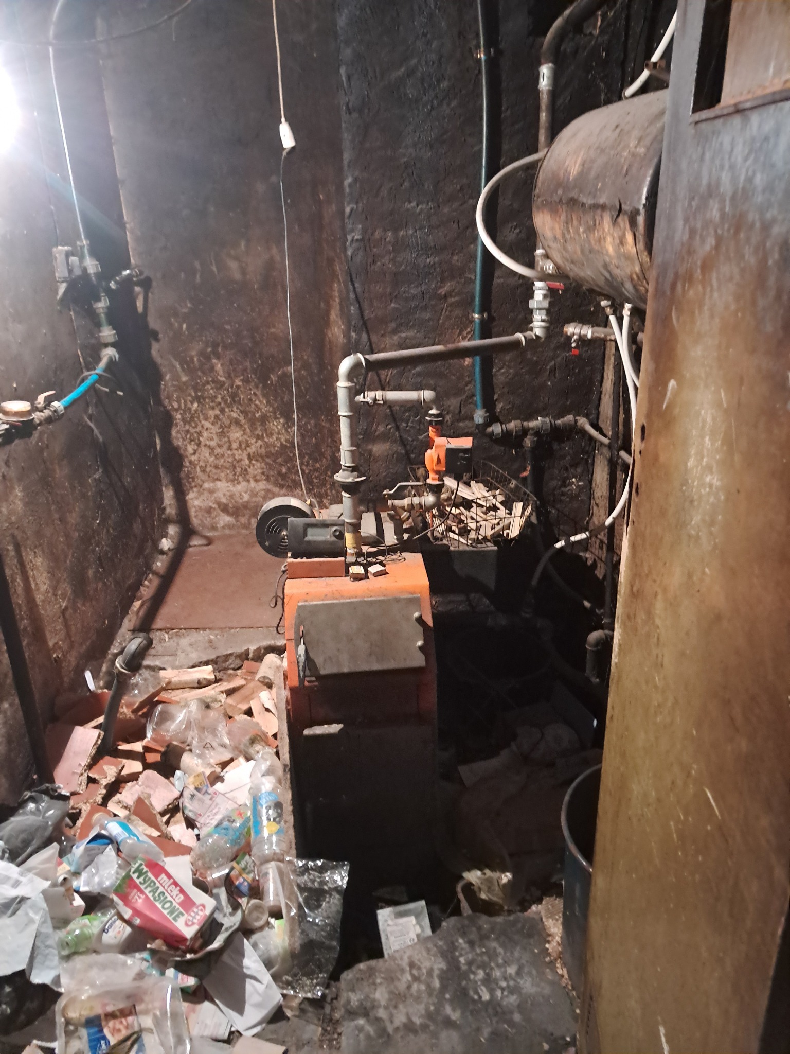 Stara kotłownia z piecem centralnego ogrzewania, widoczne rury, elementy instalacji i śmieci na podłodze w ciemnym pomieszczeniu.