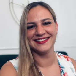 Uśmiechnięta kobieta z blond włosami i czerwonymi ustami, ubrana w bluzkę w kwiaty, na tle białej ściany z dekoracją.